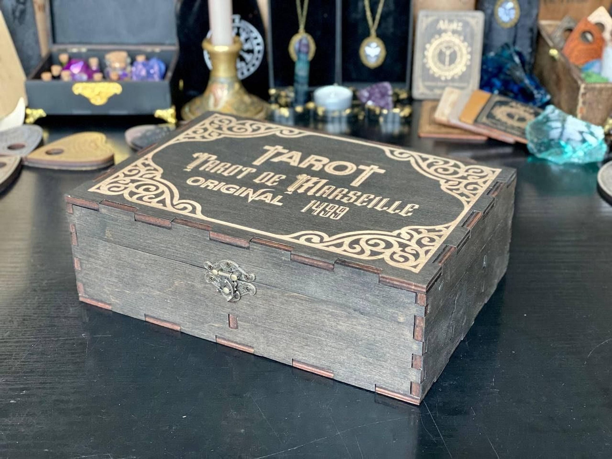 A wooden box with a decorative border, labeled "TAROT DE MANSEILLE", sits on a table surrounded by various objects, including crystals, books, and other items.
