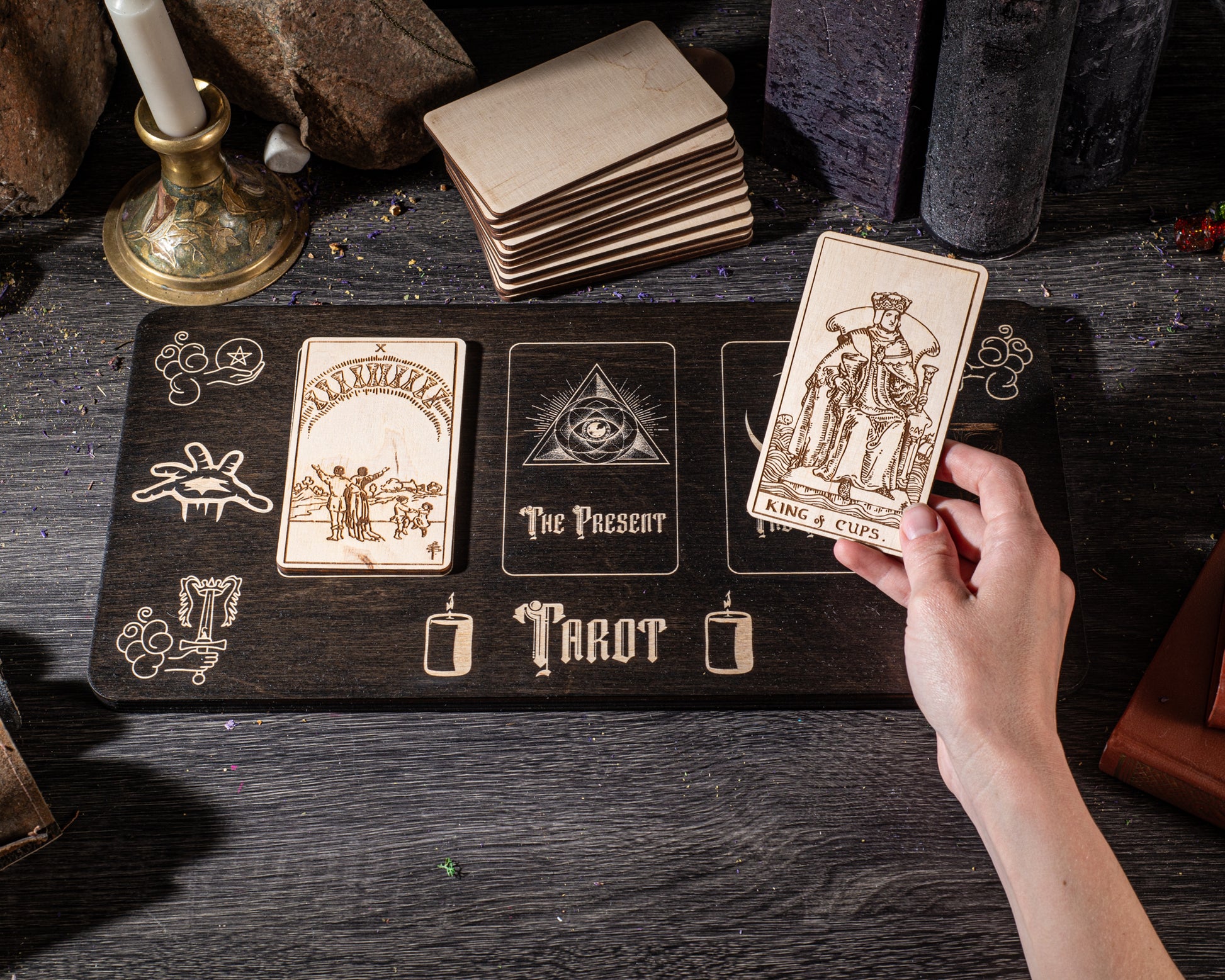 A hand is holding a tarot card over a wooden board with various other cards and objects on it, including a candle, a stack of cards, and some other items.