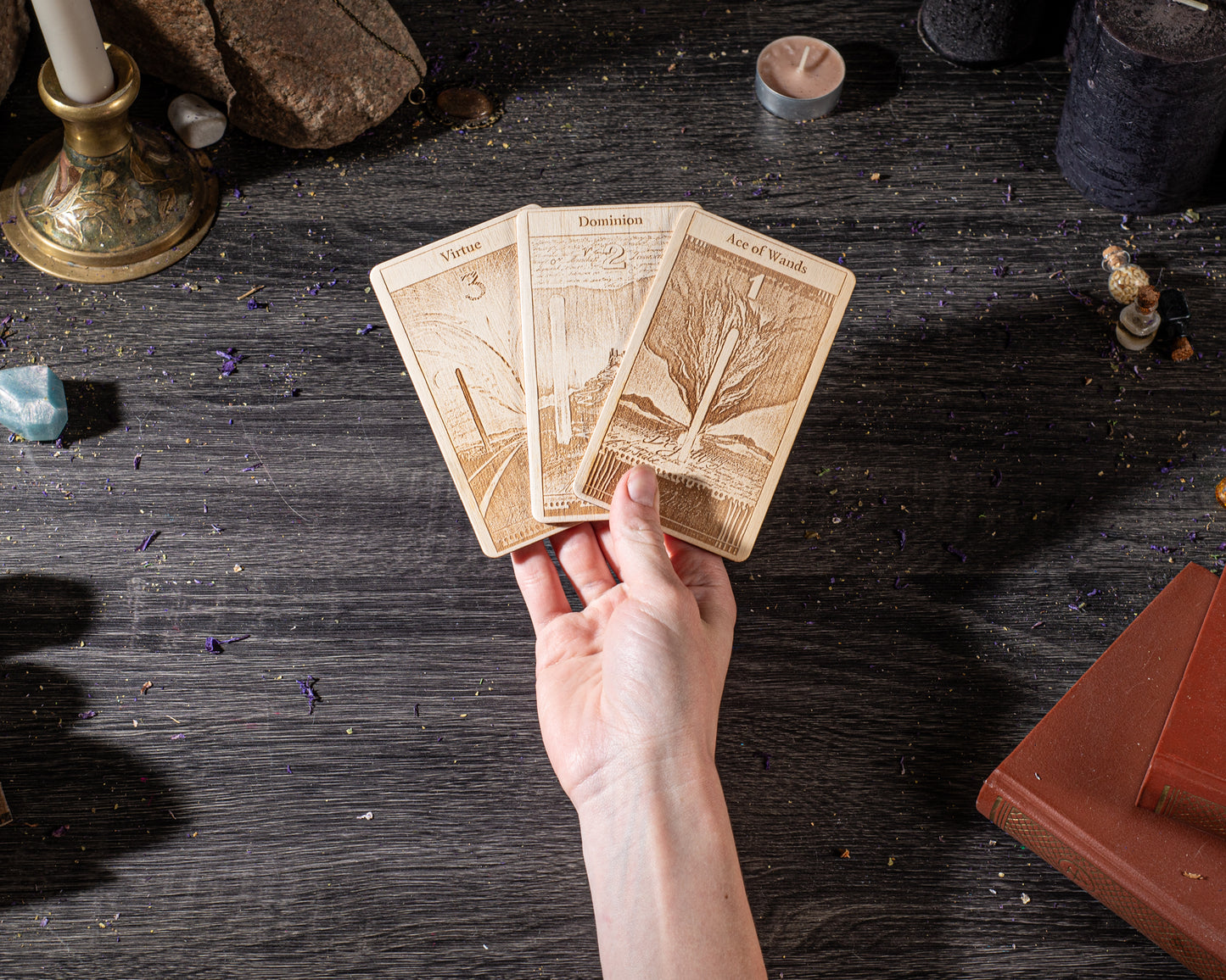 A hand holding three wooden cards with illustrations on a dark wooden surface, surrounded by various objects such as candles, crystals, and books.