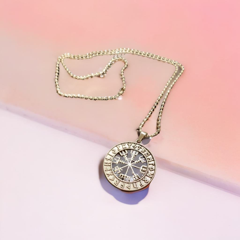A silver pendant necklace with a compass design is shown against a pink background.