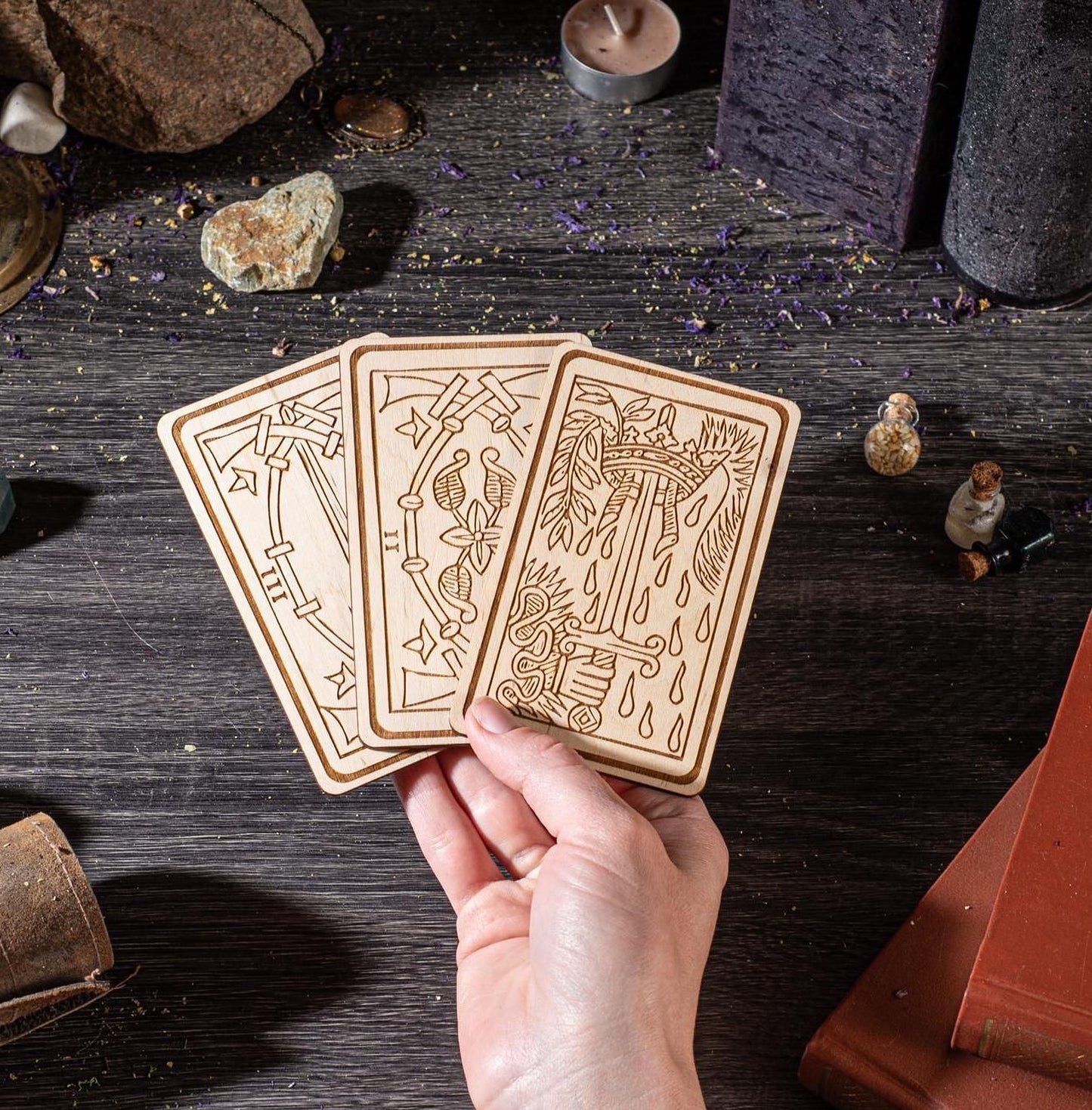 A hand is holding three wooden cards with intricate designs on a dark wooden surface, surrounded by various objects such as rocks, candles, and figurines.