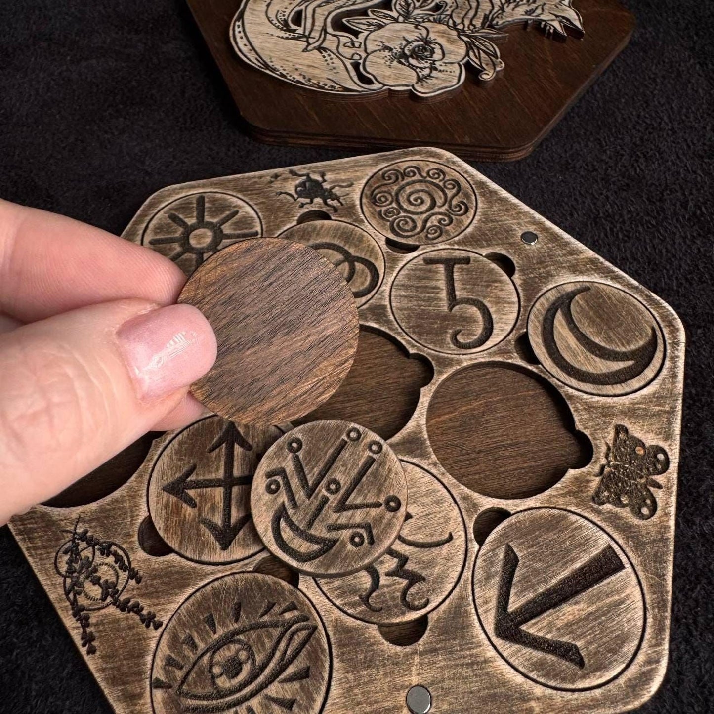 A hand is holding a wooden piece with various engraved symbols and designs, including a skull and crossbones, a crescent moon, and other abstract shapes.