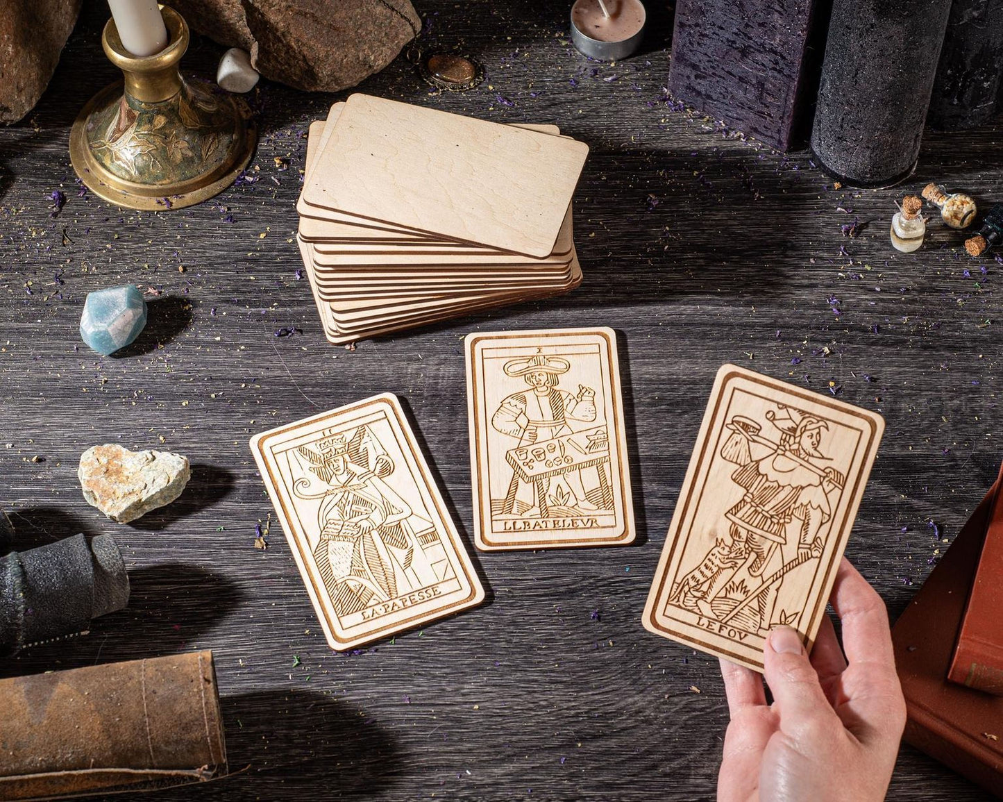 a hand holding a wooden tarot card, surrounded by various objects such as candles, crystals, and other items on a wooden surface.